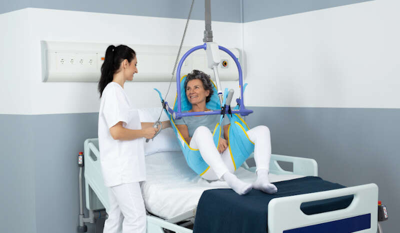 A senior woman in a Maxi Sky 2 above her bed with her nurse beside her. 