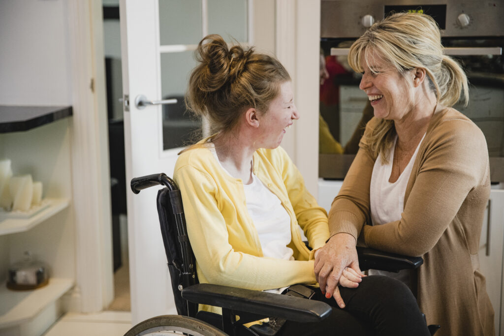 Mother with hands on lap of daughter seated in a manual wheelchair looking and smiling at each other. 