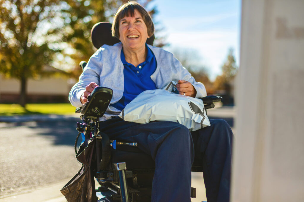Smiling senior woman outdoors on a sunny day using a power wheelchair and holding a plastic package on her lap. 