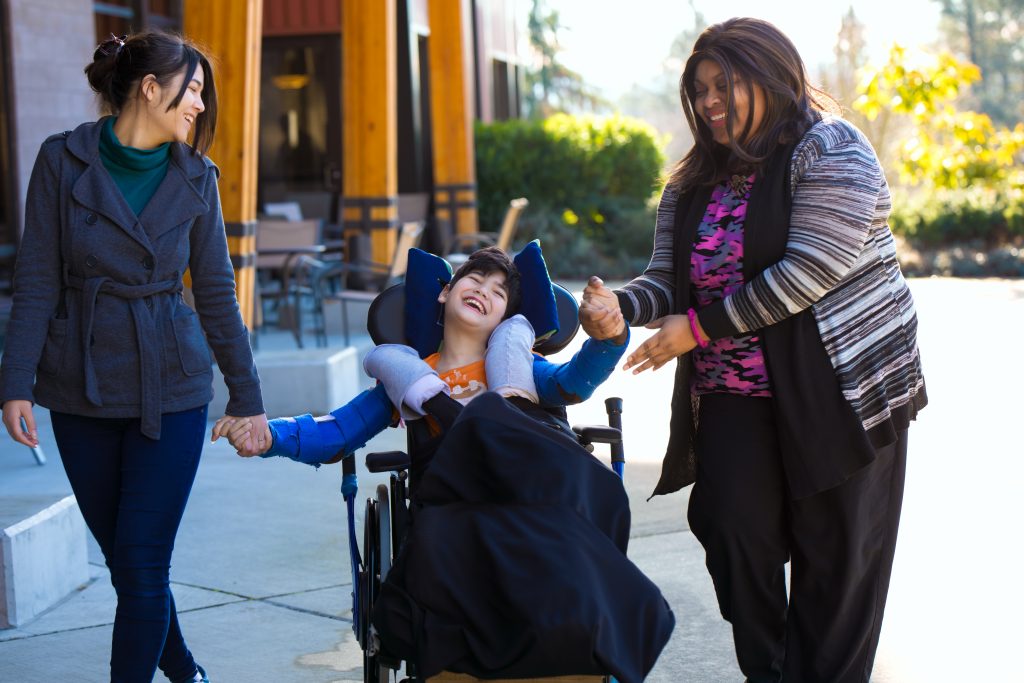 Young boy in wheelchair with 2 woman caregivers walking on a street in town