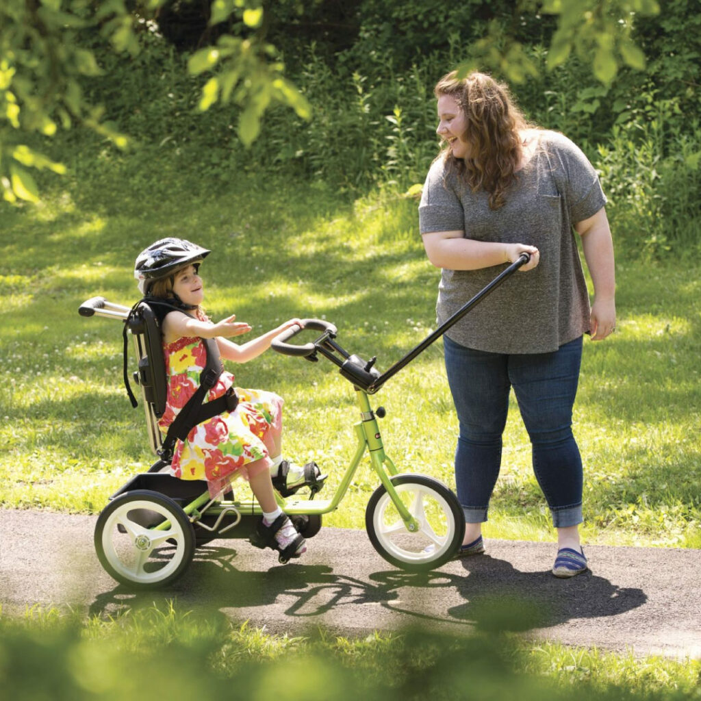 Young girl in tricycle assisted by woman on park trail.