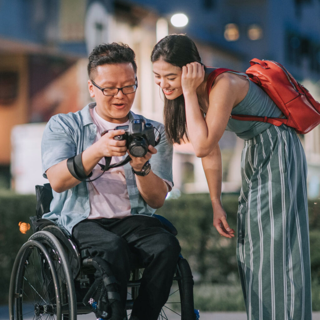 Young man in a manual wheelchair showing an image on his digital camera to a young woman standing on his left side.