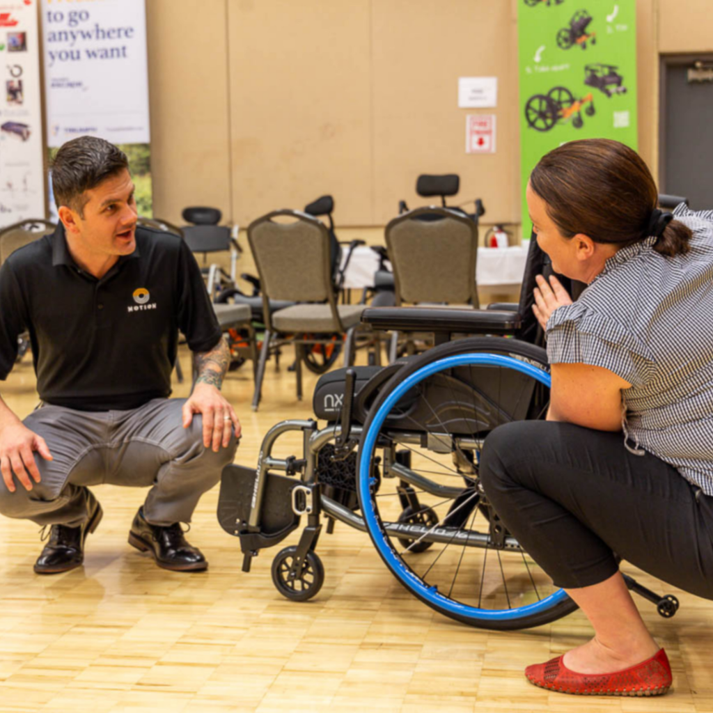 Male Motion employee kneeling down beside manual wheelchair discussing alongside kneeling woman